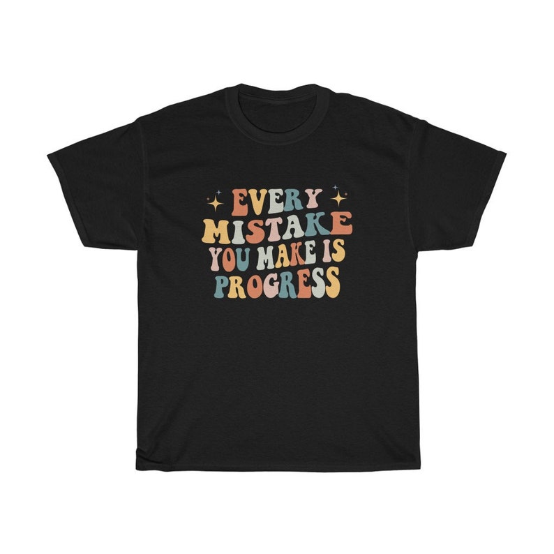 May include: Black t-shirt with the text "EVERY MISTAKE YOU MAKE IS PROGRESS" in colorful, retro-style lettering. The design includes stars and a vintage font, perfect for casual wear.