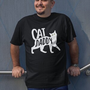 May include: Black t-shirt with the words "CAT DADDY" in white, accompanied by a white cat silhouette. The shirt is worn by a person in blue jeans. The person is smiling and leaning against a gray wall.