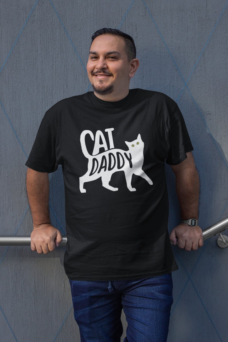 May include: Black t-shirt with the words "CAT DADDY" in white, accompanied by a white cat silhouette. The shirt is worn by a person in blue jeans. The person is smiling and leaning against a gray wall.