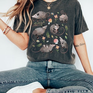 May include: A dark gray t-shirt with a graphic print of multiple opossums in a forest setting. The opossums are surrounded by flowers, leaves, and mushrooms.