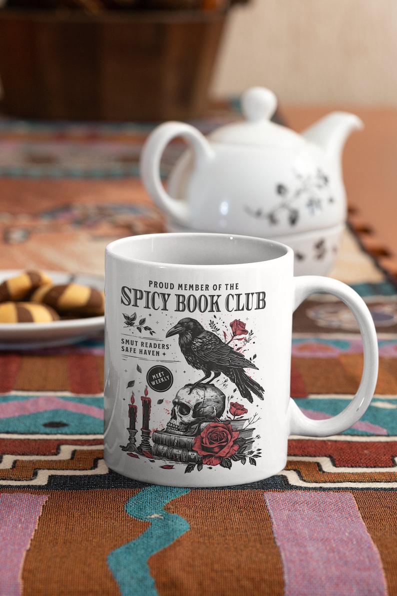 Puede incluir: Taza de cer&aacute;mica blanca con una ilustraci&oacute;n en blanco y negro de un cuervo posado sobre un cr&aacute;neo con una rosa roja. La taza dice "Proud Member of the Spicy Book Club" y "Smut Readers' Safe Haven + Meet Weekly".
