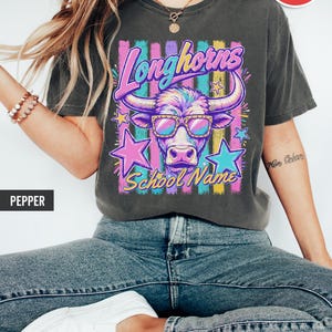 May include: A pepper-colored short-sleeve t-shirt featuring a graphic of a longhorn wearing sunglasses. The design includes the text Longhorns and School Name, surrounded by colorful vertical stripes and stars in pink, blue, and yellow hues.