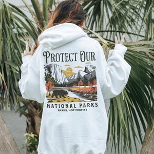 May include: White hoodie with a graphic on the back that says "PROTECT OUR NATIONAL PARKS" with a landscape illustration of a bear, river, and mountains. The text "PARKS, NOT PROFITS" is below the illustration.