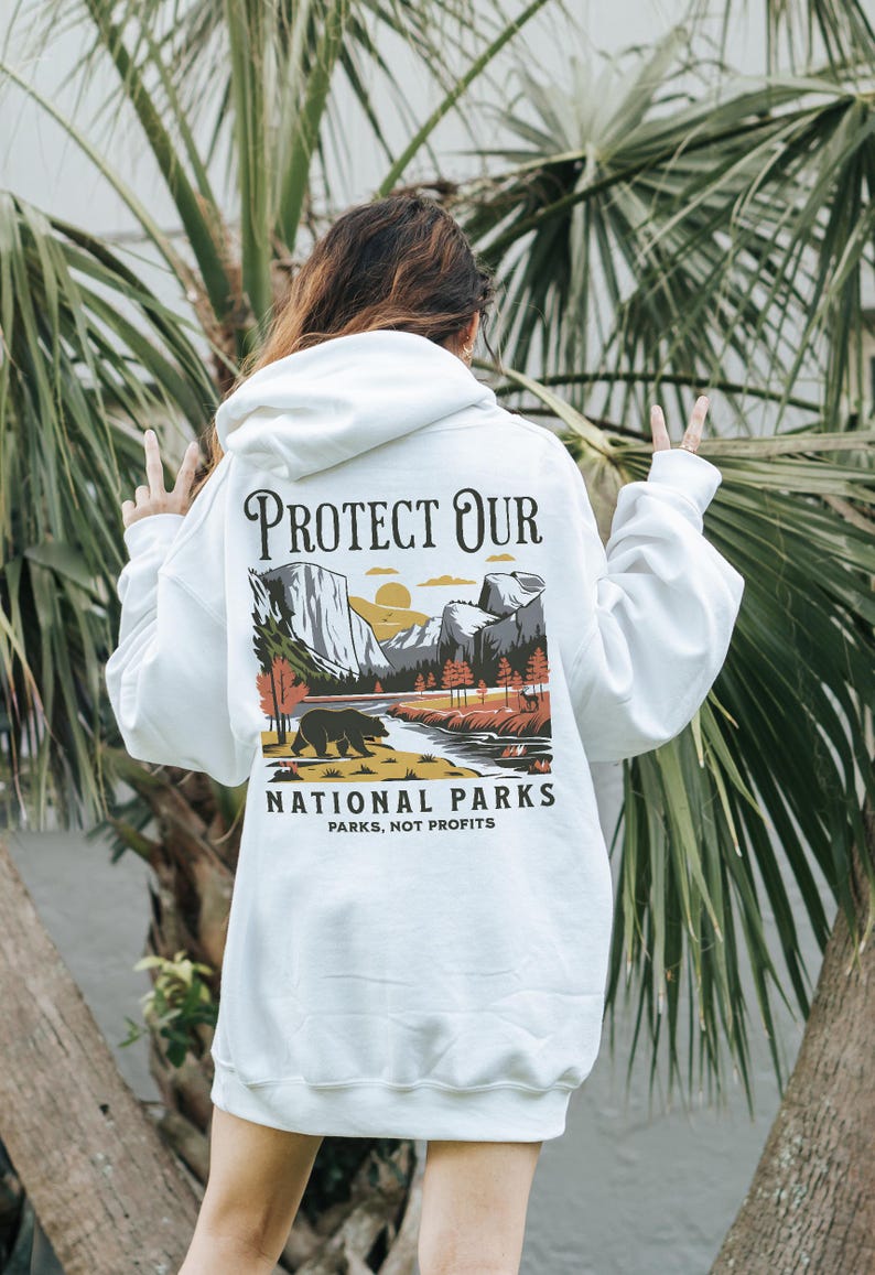 May include: White hoodie with a graphic on the back that says "PROTECT OUR NATIONAL PARKS" with a landscape illustration of a bear, river, and mountains. The text "PARKS, NOT PROFITS" is below the illustration.