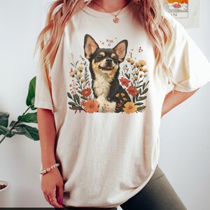 K&ouml;nnte beinhalten: Ein wei&szlig;es T-Shirt mit einem floralen Design, das einen Chihuahua zeigt. Der Text "MODEL IS WEARING A SIZE XL COLOR: IVORY" befindet sich am unteren Rand des Bildes.