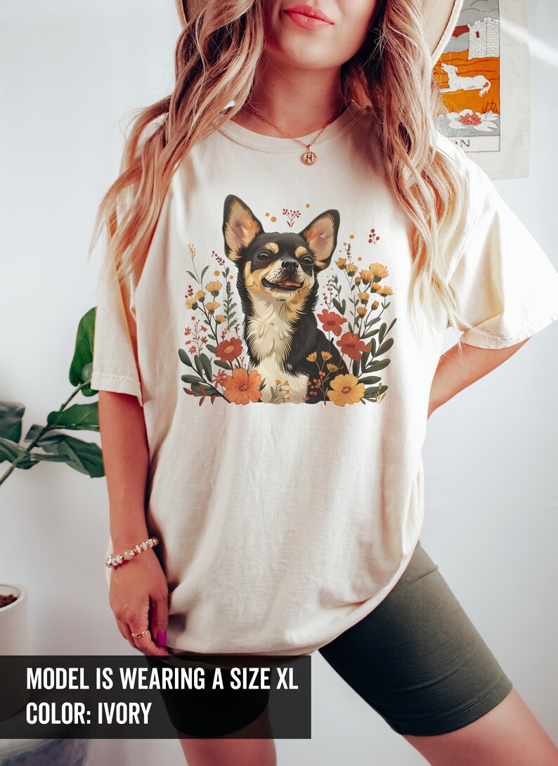K&ouml;nnte beinhalten: Ein wei&szlig;es T-Shirt mit einem floralen Design, das einen Chihuahua zeigt. Der Text "MODEL IS WEARING A SIZE XL COLOR: IVORY" befindet sich am unteren Rand des Bildes.