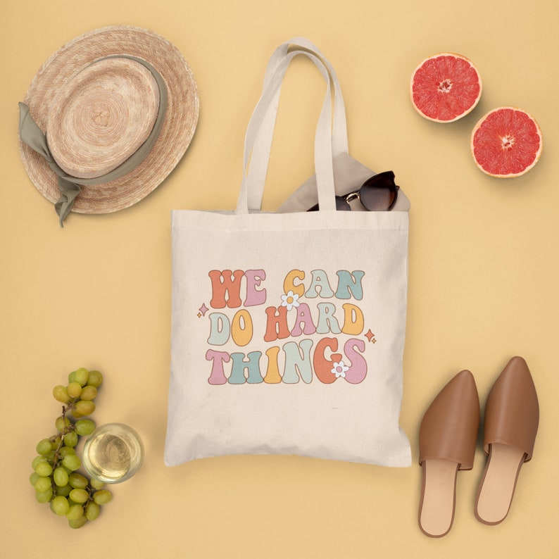 Op de afbeelding: Een witte canvas tote bag met de tekst "WE CAN DO HARD THINGS" in kleurrijke retro letters met kleine bloem accenten. De tas is op een gele achtergrond met andere items zoals een hoed, zonnebril en fruit.