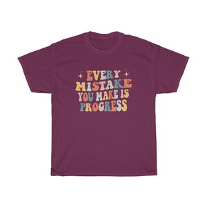 May include: Burgundy t-shirt with the text "EVERY MISTAKE YOU MAKE IS PROGRESS" in a retro font. The text is in various colors, including orange, blue, and yellow. The shirt has a crew neck and short sleeves.