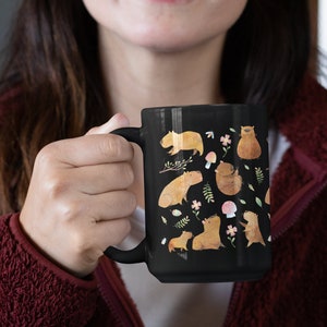 May include: A black ceramic mug with a white handle features a whimsical design of capybaras, flowers, and mushrooms. The capybaras are depicted in various poses, including sitting, standing, and swimming.