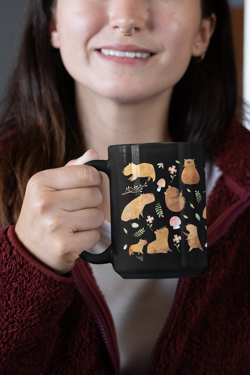 May include: A black ceramic mug with a white handle features a whimsical design of capybaras, flowers, and mushrooms. The capybaras are depicted in various poses, including sitting, standing, and swimming.