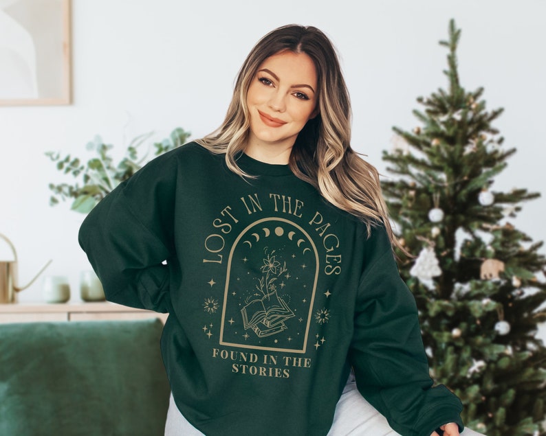 May include: A dark green sweatshirt with a gold graphic design featuring a book, moon phases, and stars. The text on the sweatshirt reads "Lost in the Pages Found in the Stories".