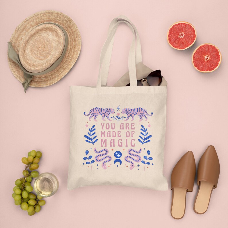 Op de afbeelding: Cr&egrave;mekleurige draagtas met een ontwerp met twee roze tijgers, blauwe bladeren en de tekst "YOU ARE MADE OF MAGIC". De tas heeft lange handvatten en is gemaakt van een stevige stof. De tas staat op een roze achtergrond.