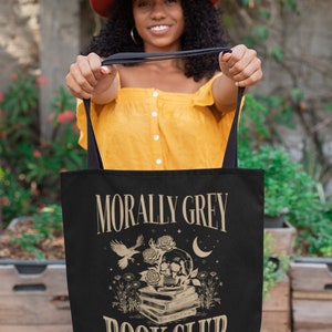 May include: A black tote bag with a white design that says "Morally Grey Book Club" with a skull and crossbones, a bird, and a crescent moon. The text below the main design says "Enemies to Lovers Weekly".