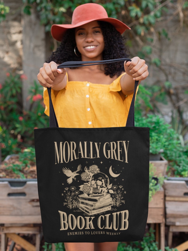 May include: A black tote bag with a white design that says "Morally Grey Book Club" with a skull and crossbones, a bird, and a crescent moon. The text below the main design says "Enemies to Lovers Weekly".