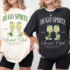May include: Two t-shirts with a graphic design featuring a cocktail drink with the text "Hugo Spritz" and "Social Club" with the dates "MAR 2" and "MAR 2025". The t-shirts are available in ivory and black.