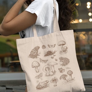 May include: A beige tote bag with a brown illustration of mushrooms, frogs, butterflies, and other forest elements. The bag has two long straps.