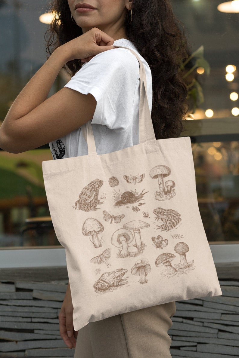 May include: A beige tote bag with a brown illustration of mushrooms, frogs, butterflies, and other forest elements. The bag has two long straps.