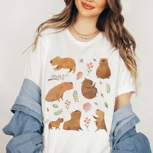 May include: White t-shirt with a colorful illustration of capybaras in various poses, surrounded by flowers, leaves, and mushrooms.
