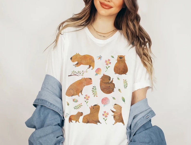 May include: White t-shirt with a colorful illustration of capybaras in various poses, surrounded by flowers, leaves, and mushrooms.