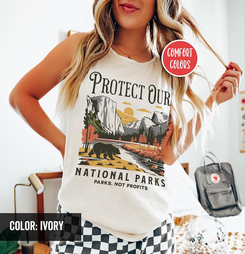 May include: Ivory-colored tank top with a graphic featuring a bear, mountains, and the text "Protect Our National Parks, Parks Not Profits." The design includes a sunset and trees. The shirt is labeled "Comfort Colors."