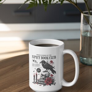 Puede incluir: Taza de cer&aacute;mica blanca con una ilustraci&oacute;n en blanco y negro de un cuervo posado sobre un cr&aacute;neo con una pila de libros y velas. El texto dice "Proud Member of the Spicy Book Club" y "Smut Readers Safe Haven + Meet Weekly".