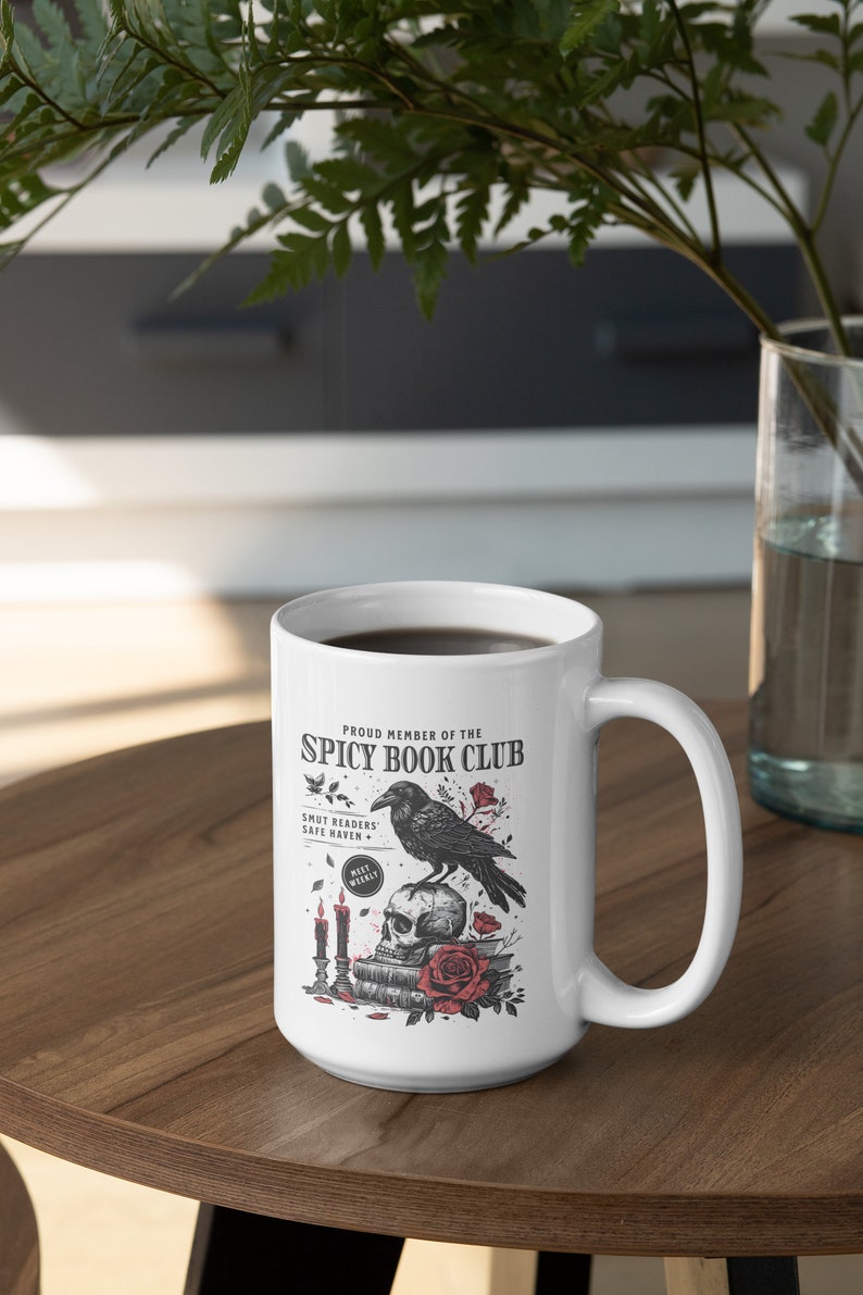 Puede incluir: Taza de cer&aacute;mica blanca con una ilustraci&oacute;n en blanco y negro de un cuervo posado sobre un cr&aacute;neo con una pila de libros y velas. El texto dice "Proud Member of the Spicy Book Club" y "Smut Readers Safe Haven + Meet Weekly".