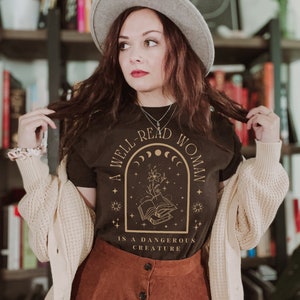 K&ouml;nnte beinhalten: Ein dunkelbraunes T-Shirt mit goldenem Text "A Well-Read Woman Is A Dangerous Creature". Das Design zeigt ein florales und himmlisches Motiv. Die Person tr&auml;gt einen grauen Hut, eine cremefarbene Strickjacke und einen braunen Rock.