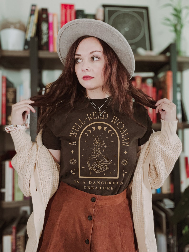 K&ouml;nnte beinhalten: Ein dunkelbraunes T-Shirt mit goldenem Text "A Well-Read Woman Is A Dangerous Creature". Das Design zeigt ein florales und himmlisches Motiv. Die Person tr&auml;gt einen grauen Hut, eine cremefarbene Strickjacke und einen braunen Rock.
