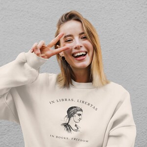 May include: A white sweatshirt with a black graphic print of a woman's head with a laurel wreath and the text "In Libras, Libertas" and "In Books, Freedom".