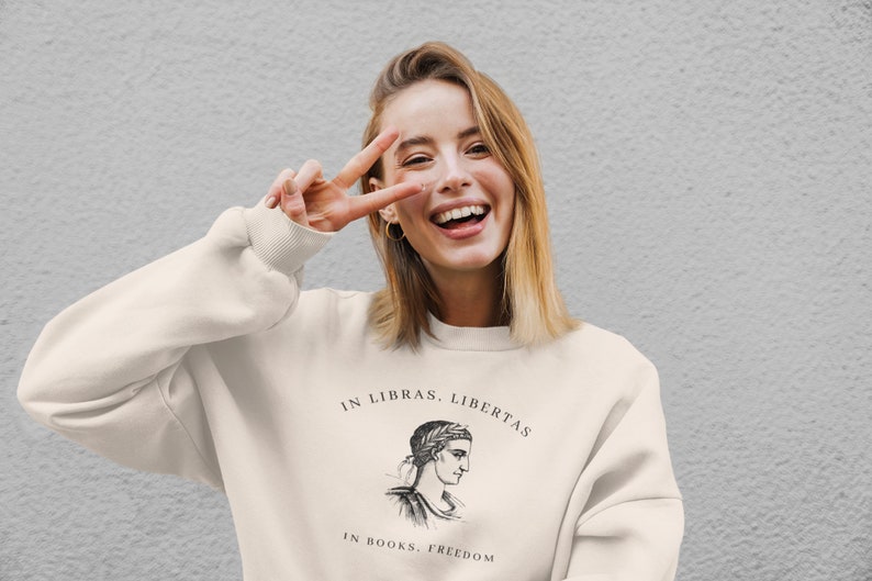 May include: A white sweatshirt with a black graphic print of a woman's head with a laurel wreath and the text "In Libras, Libertas" and "In Books, Freedom".