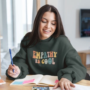 May include: A dark green sweatshirt with the text "EMPATHY IS COOL" in rainbow colors and smiley faces. The sweatshirt is worn by a person sitting at a desk with a notebook and pen.