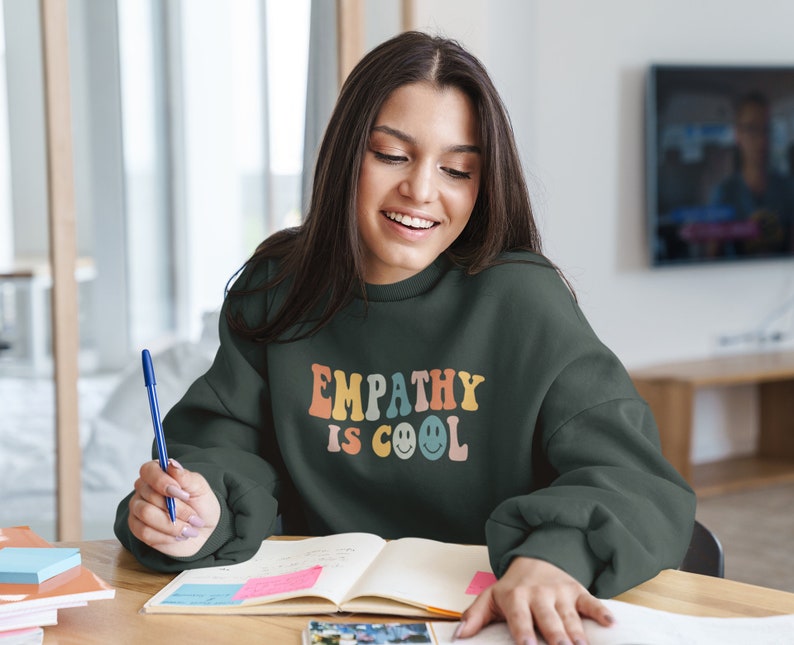 May include: A dark green sweatshirt with the text "EMPATHY IS COOL" in rainbow colors and smiley faces. The sweatshirt is worn by a person sitting at a desk with a notebook and pen.