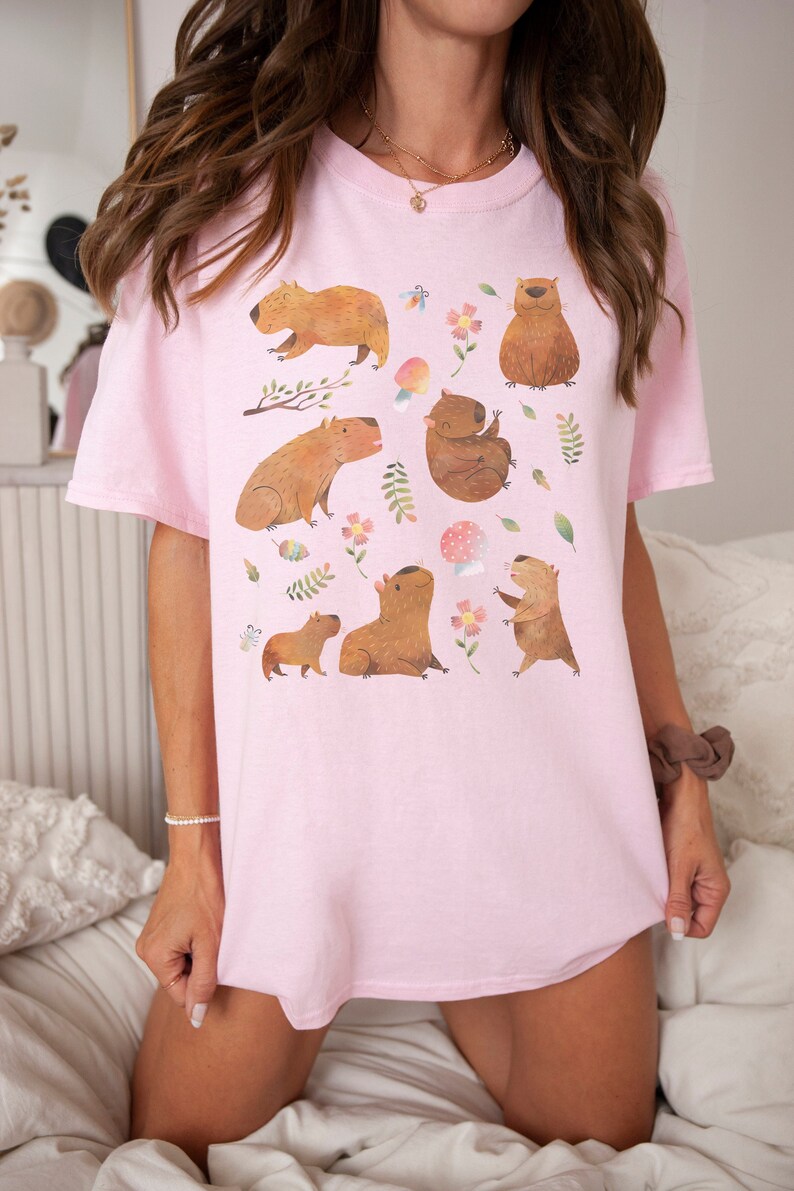 May include: A pink oversized t-shirt with a graphic print of multiple capybaras in a forest setting. The capybaras are brown and are depicted in various poses, including sitting, standing, and walking. The forest setting includes green leaves, pink flowers, and a mushroom.