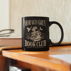 May include: Black coffee mug with a white design. The design features a skull with roses, a bird, and a crescent moon. The text reads "Morally Grey Book Club Enemies to Lovers Weekly".