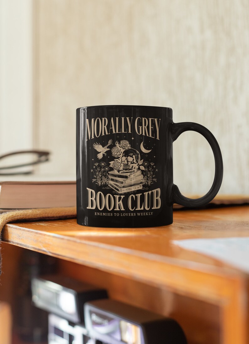 May include: Black coffee mug with a white design. The design features a skull with roses, a bird, and a crescent moon. The text reads "Morally Grey Book Club Enemies to Lovers Weekly".