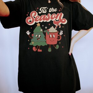 May include: Black t-shirt with a retro-style graphic of a Christmas tree and a mug with the text "Tis' the Season".