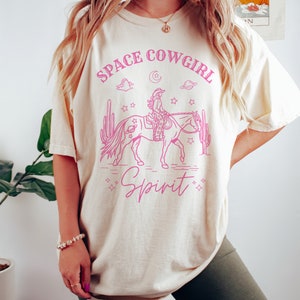 May include: A cream-colored t-shirt with a pink graphic of a cowgirl riding a horse in space. The graphic includes the text "Space Cowgirl Spirit".