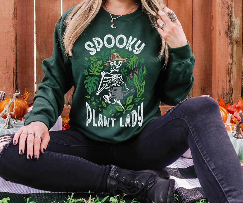 May include: A dark green sweatshirt with a white graphic of a skeleton wearing a hat and holding a watering can. The graphic says "Spooky Plant Lady".