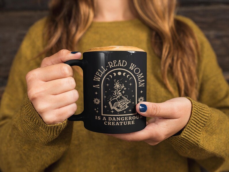 May include: Black ceramic mug with a handle, filled with a light-colored beverage. The mug features the text "A WELL-READ WOMAN IS A DANGEROUS CREATURE" in a decorative design with moons, stars, and an open book.