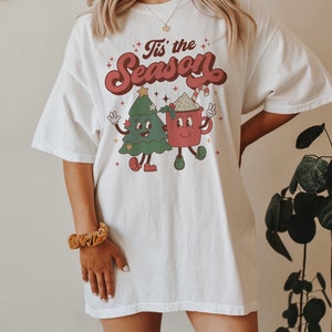 May include: White oversized t-shirt with a retro-style graphic of a Christmas tree and a mug of hot chocolate. The text "Tis' the Season" is printed in a festive font above the graphic.