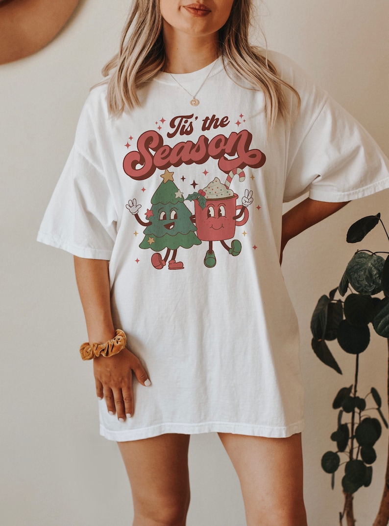 May include: White oversized t-shirt with a retro-style graphic of a Christmas tree and a mug of hot chocolate. The text "Tis' the Season" is printed in a festive font above the graphic.