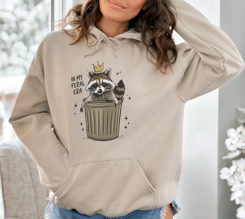 May include: A beige hoodie featuring a graphic of a raccoon wearing a gold crown and sitting in a bin. The text "In my feral era" is printed on the hoodie.