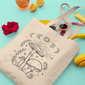 K&ouml;nnte beinhalten: Eine beige Tragetasche mit einer schwarz-wei&szlig;en Illustration von Pilzen und einem Halbmond-Design. Die Tasche hat lange Henkel und liegt auf einer t&uuml;rkisfarbenen Oberfl&auml;che.