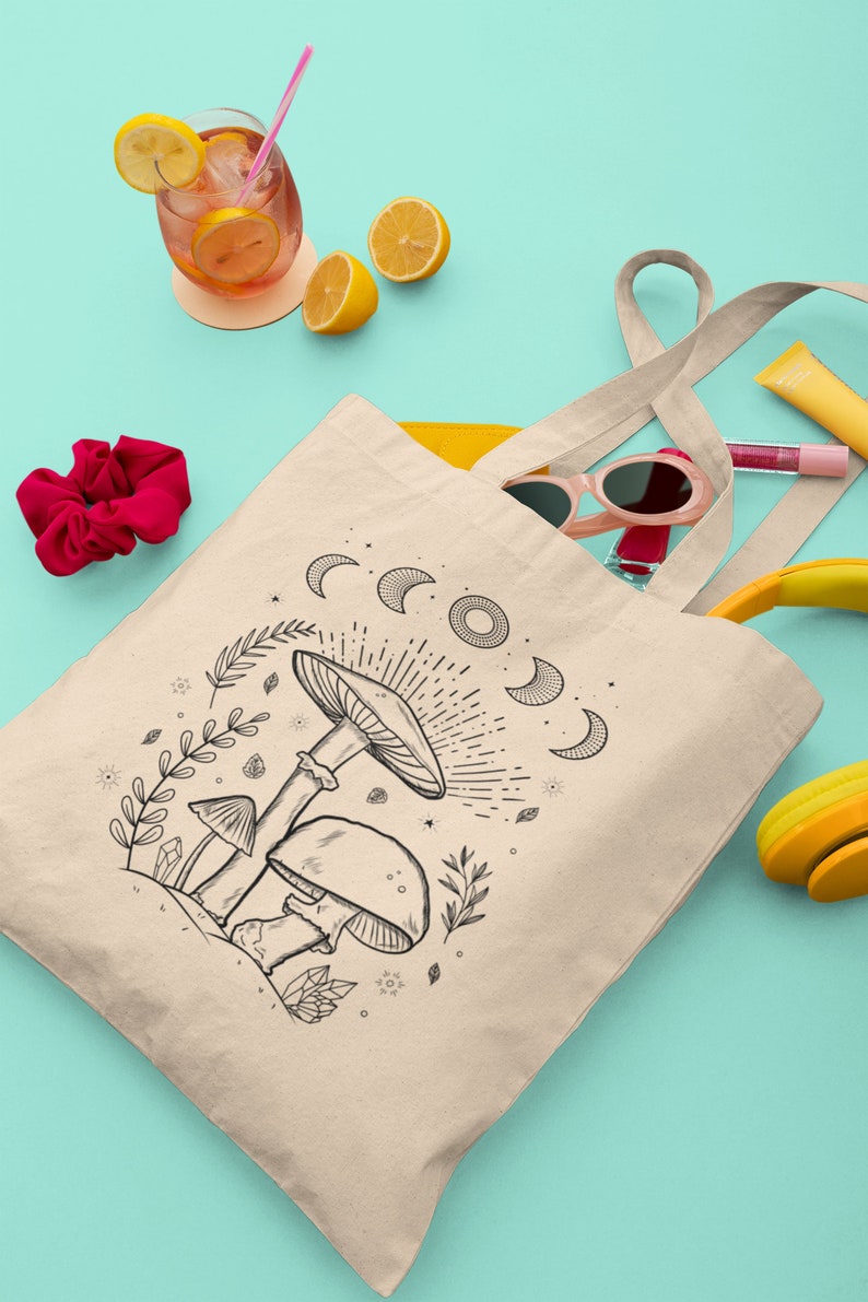 K&ouml;nnte beinhalten: Eine beige Tragetasche mit einer schwarz-wei&szlig;en Illustration von Pilzen und einem Halbmond-Design. Die Tasche hat lange Henkel und liegt auf einer t&uuml;rkisfarbenen Oberfl&auml;che.
