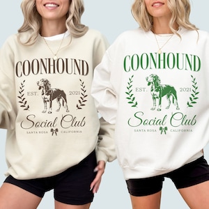 May include: Two white sweatshirts with a brown and green graphic design. The design features a dog, the words "Coonhound Social Club", and the text "Est. 2021".