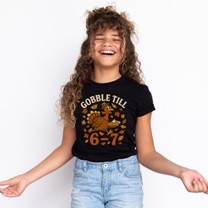 K&ouml;nnte beinhalten: Schwarzes T-Shirt mit dem Text "GOBBLE TILL" &uuml;ber einer Cartoon-Truthahn, der einen Kuchen h&auml;lt, umgeben von Herbstbl&auml;ttern. Der Text "6-7" steht unter dem Truthahn. Das T-Shirt wird von einer Person getragen, die eine blaue Jeans tr&auml;gt.