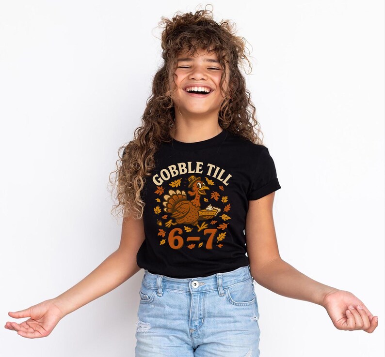 K&ouml;nnte beinhalten: Schwarzes T-Shirt mit dem Text "GOBBLE TILL" &uuml;ber einer Cartoon-Truthahn, der einen Kuchen h&auml;lt, umgeben von Herbstbl&auml;ttern. Der Text "6-7" steht unter dem Truthahn. Das T-Shirt wird von einer Person getragen, die eine blaue Jeans tr&auml;gt.