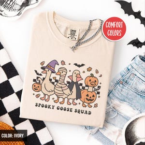 May include: Ivory t-shirt with a Halloween design of four cartoon geese in costumes: a witch's hat, mummy wrap, cape, and pumpkin. The text "SPOOKY GOOSE SQUAD" is below. The shirt has a V-neck and a silver chain. The shirt is ivory in colour.