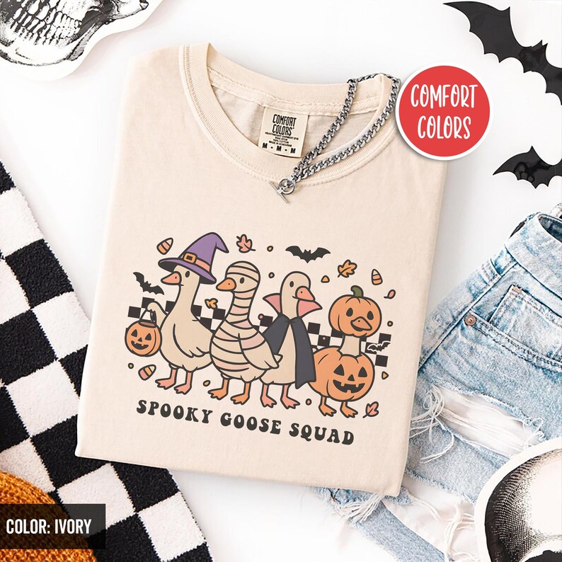 May include: Ivory t-shirt with a Halloween design of four cartoon geese in costumes: a witch's hat, mummy wrap, cape, and pumpkin. The text "SPOOKY GOOSE SQUAD" is below. The shirt has a V-neck and a silver chain. The shirt is ivory in colour.