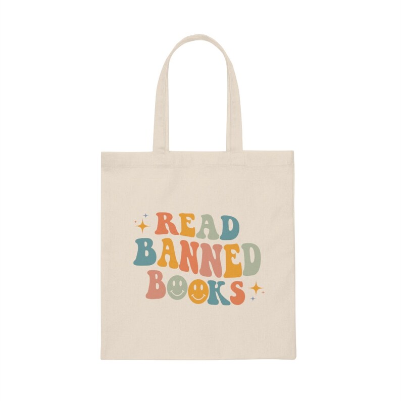 Peut inclure: Un sac fourre-tout beige avec le texte "READ BANNED BOOKS" dans un arc-en-ciel de couleurs. Le texte est dans une police r&eacute;tro avec un smiley au milieu du mot "BOOKS".